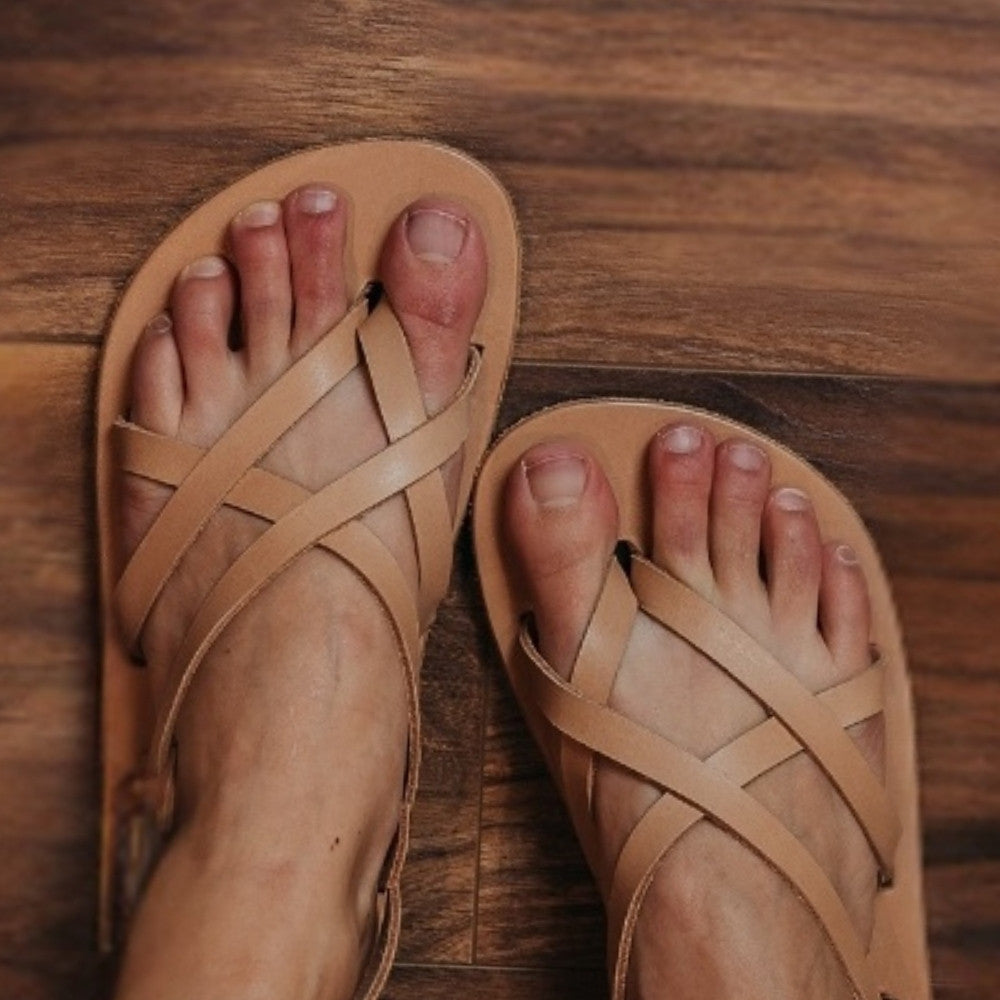 Tan, leather, strappy sandal with a silver buckle in the back. Both sandals are shown from above on a light-skinned woman sitting on a brown wooden floor. #color_tan