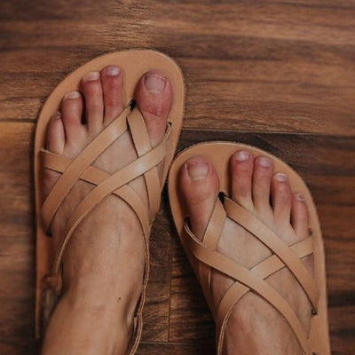 Tan, leather, strappy sandal with a silver buckle in the back. Both sandals are shown from above on a light-skinned woman sitting on a brown wooden floor. #color_tan