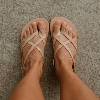 Tan, leather, strappy sandal with a silver buckle in the back. Both sandals are shown from above on a tan woman standing on cement. #color_tan