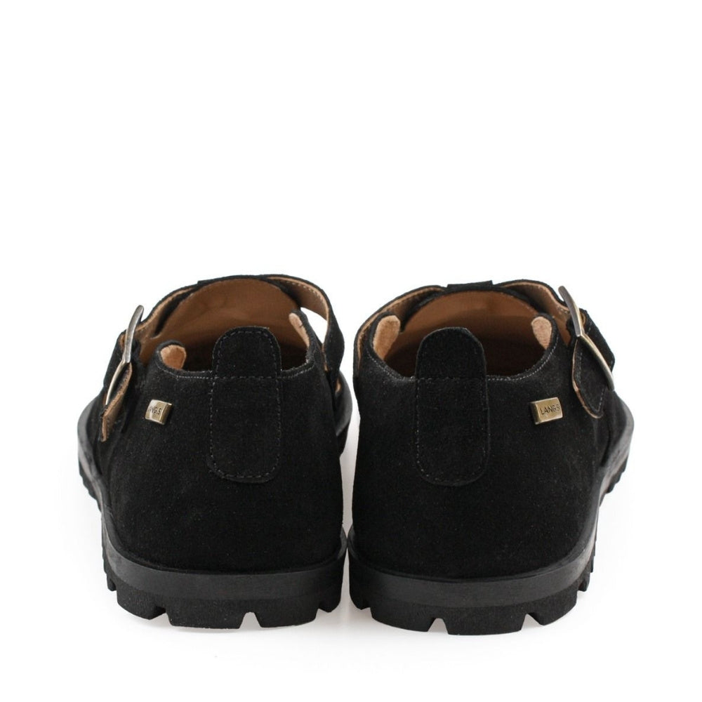 A black, suede t-bar flat with tan lining, a gold buckle, thick black soles, and a small pull tab in the back. Both shoes are shown from the back against a white background. #color_black