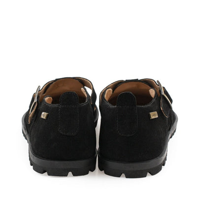 A black, suede t-bar flat with tan lining, a gold buckle, thick black soles, and a small pull tab in the back. Both shoes are shown from the back against a white background. #color_black
