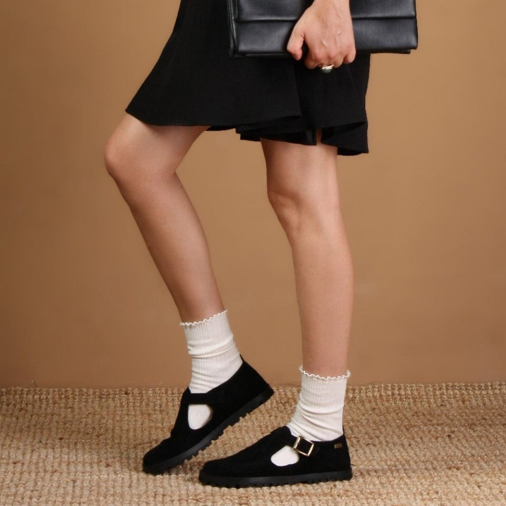 A black, suede t-bar flat with tan lining, a gold buckle, thick black soles, and a small pull tab in the back. Both shoes are shown facing left on a light-skinned woman standing on a white jute rug in a tan studio. She is wearing a short, black, pleated skirt and cream ruffle top socks holding a black, leather, envelope bag.  #color_black