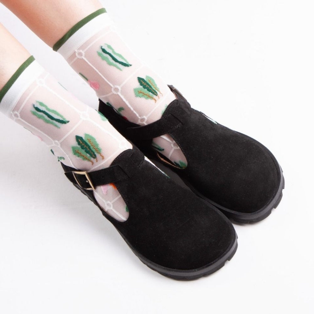 A black, suede t-bar flat with tan lining, a gold buckle, thick black soles, and a small pull tab in the back. Both shoes are shown from above facing diagonally right on a light-skinned woman sitting on a white floor. She is wearing white, see-through socks with a quilted foliage design.  #color_black