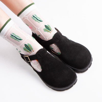 A black, suede t-bar flat with tan lining, a gold buckle, thick black soles, and a small pull tab in the back. Both shoes are shown from above facing diagonally right on a light-skinned woman sitting on a white floor. She is wearing white, see-through socks with a quilted foliage design.  #color_black