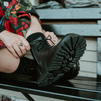 Black, smooth leather Lang.S Kombat boot, with round black laces, a pull tab, zipper, and chinky black soles. Both shoes are shown on a light-skinned woman sitting on a black bench in an urban setting with her legs crossed wearing a colorful, black floral jumpsuit. #color_black