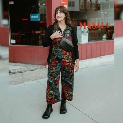 Black, smooth leather Lang.S Kombat boot, with round black laces, a pull tab, zipper, and chinky black soles. Both shoes are shown from the front on a light-skinned woman with short, curly brown hair with bangs standing on a sidewalk in an urban setting. She is wearing a colorful, black floral jumpsuit, a black sweater, and a black, leather cross-body bag. #color_black