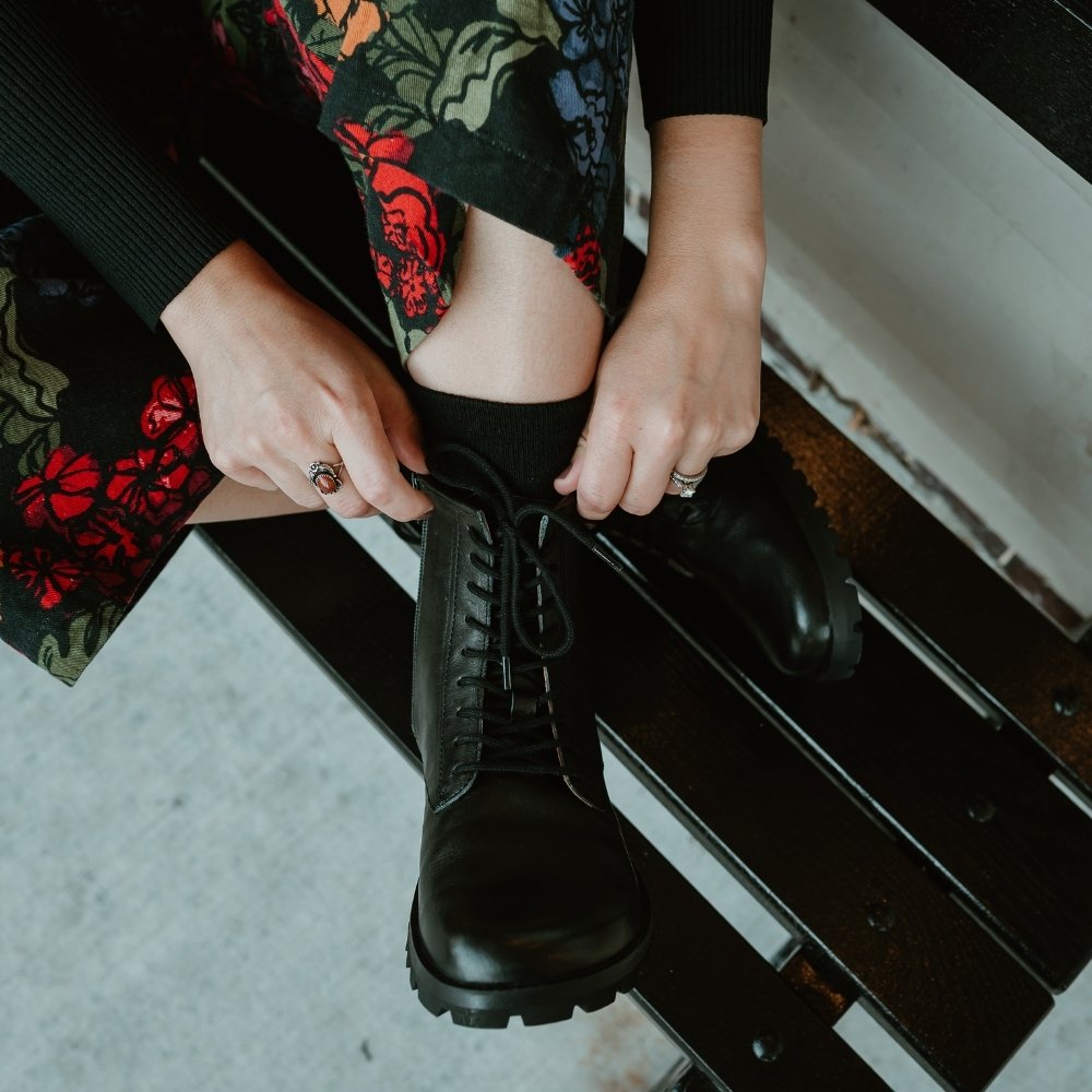 Black, smooth leather Lang.S Kombat boot, with round black laces, a pull tab, zipper, and chinky black soles. Both shoes are shown from above on a light-skinned woman sitting on a black bench in an urban setting with her legs crossed wearing a colorful, black floral jumpsuit. #color_black