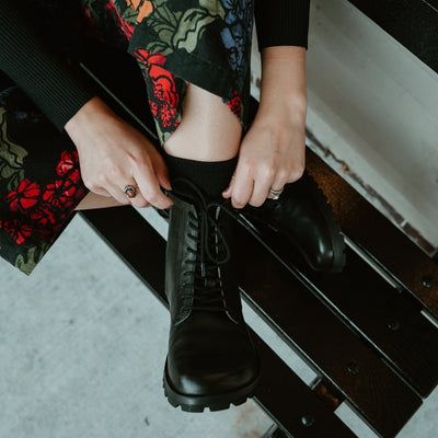 Black, smooth leather Lang.S Kombat boot, with round black laces, a pull tab, zipper, and chinky black soles. Both shoes are shown from above on a light-skinned woman sitting on a black bench in an urban setting with her legs crossed wearing a colorful, black floral jumpsuit. #color_black