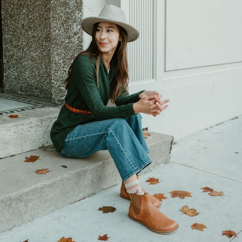 Brown leather Lang.S Luna Chelsea boots with a pull tab, tan interior leather, and tan rubber. Both boots are shown facing right on a light skinned woman wearing medium wash, loose, cropped jeans, a green sweater with a tan belt around the waist, a tan wide-brimmed hat, and tan reindeer patterned socks sitting on cement stairs. #color_brown