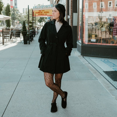 A black, suede t-bar flat with tan lining, a gold buckle, thick black soles, and a small pull tab in the back. Both shoes are shown from the front on a light-skinned woman wearing cheetah-patterned, fishnet tights, a short, black, a-line trench coat, a white short turtle-neck, and a beret standing on a cement sidewalk in an urban setting looking left.  #color_black