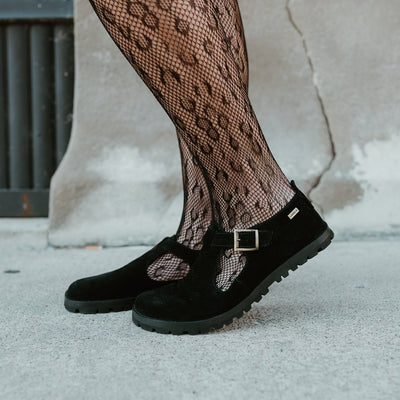 A black, suede t-bar flat with tan lining, a gold buckle, thick black soles, and a small pull tab in the back. Both shoes are shown facing left on a light-skinned woman wearing cheetah-patterned, fishnet tights standing on a cement sidewalk in an urban setting.  #color_black