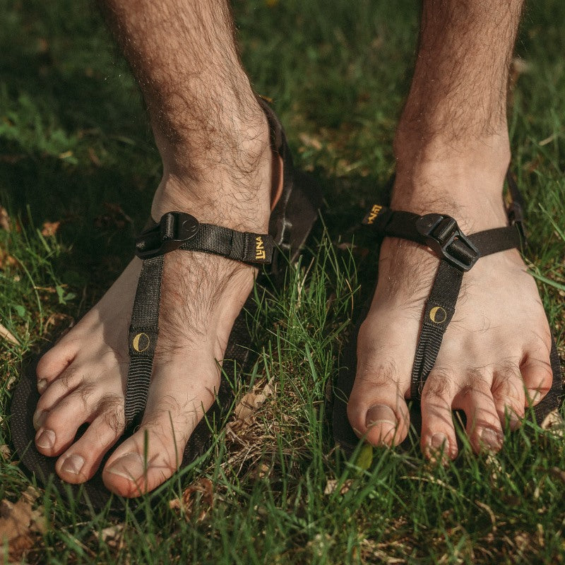 A photo of Luna Mono Winged Black Adventure Sandals made from a monkey grip technology footbed and a rubber sole. The sandals have a thong toe strap with straps that go around the ankle and heel. The sandals have a logo moon on the thong toe strap and luna logo on the heel and right ankle strap. Both sandals are shown from the front on a mans feet standing on green grass. #color_black