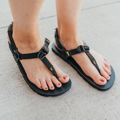 A photo of Luna Mono Winged Black Adventure Sandals made from a monkey grip technology footbed and a rubber sole. The sandals have a thong toe strap with straps that go around the ankle and heel. The sandals have a logo moon on the thong toe strap and luna logo on the heel and right ankle strap. A woman is shown from mid leg down wearing the Luna Sandals she is facing diagonally towards the right while she stands on concrete. #color_black