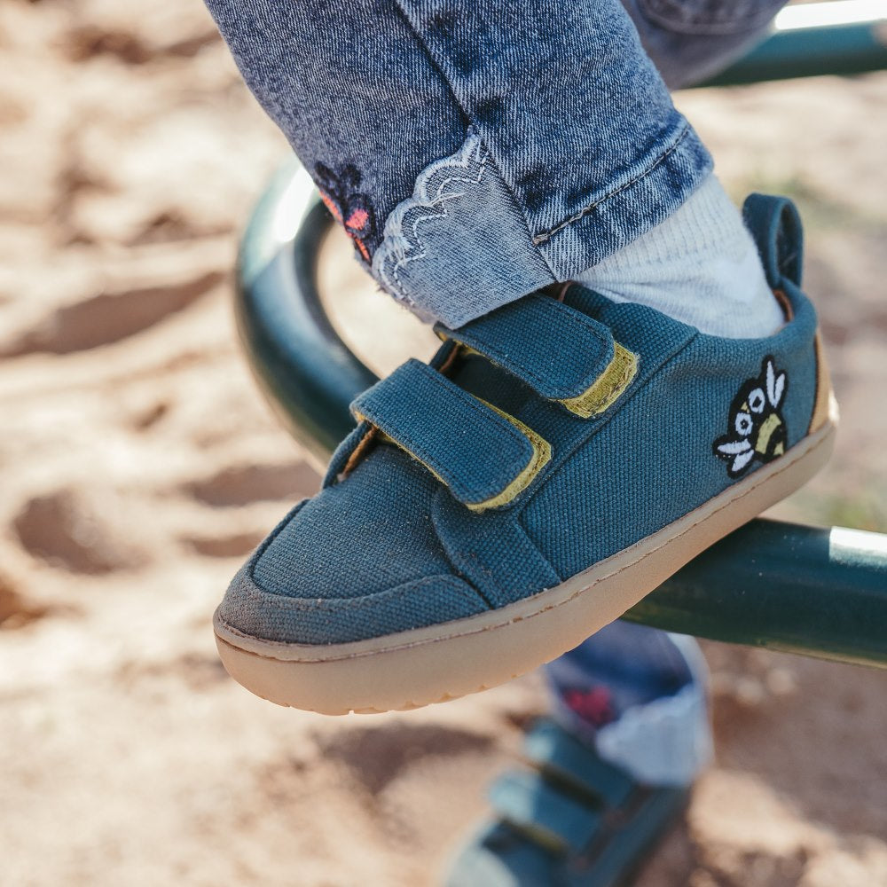 Abelha Dark Teal Mukishoes Play Kids shoe. Shoes are simple in design with velcro closures, an embroidered bee on the side close to the heel, a pull tab on the heel, a yellow color block on the heel, and yellow lining. Soles are a natural rubber gum color. Left shoe is facing left on a little girl wearing jeans stepping up onto a rounded metal playground ladder with sand in the background. #color_abelha