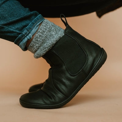 A photo of Mukishoes felted-wool-lined, leather Chelsea boots in black. Boots have elastic panels on both sides of the shoe and a pull tab at the top of the ankle opening. Soles are black in color and are stitched to the leather. The right boot is shown crouched, styled with grey socks and denim jeans, against a warm-toned background.

#color_black