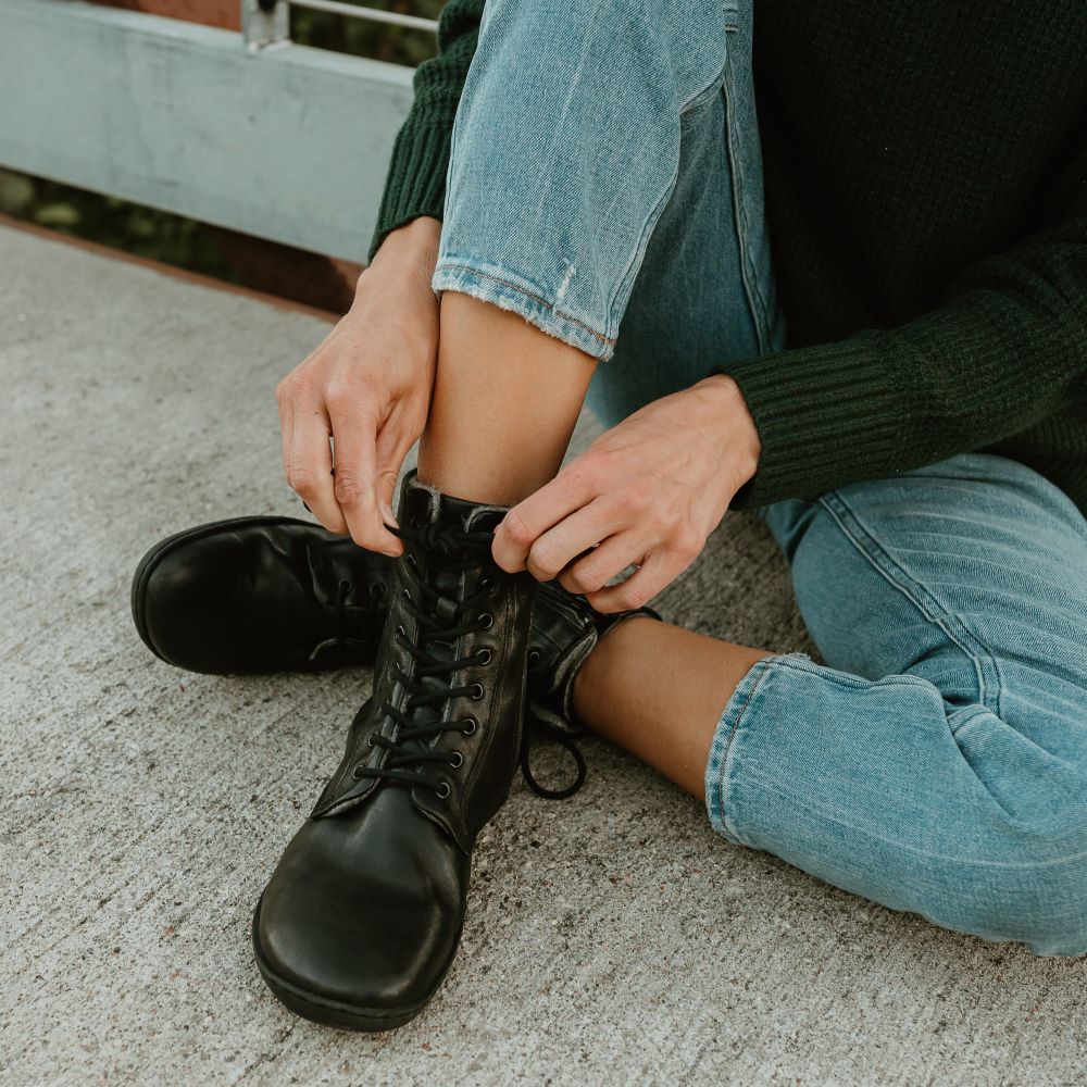 Black, leather, wool-lined Mukishoes Igneous combat-style, zip-up boots. Both boots are shown from the front on a tan woman wearing a loose black sweater and loose, cropped, light-wash jeans, sitting on a cement and metal bridge. Her right leg is crossed over the right with the right leg leaning to the side.  #color_black