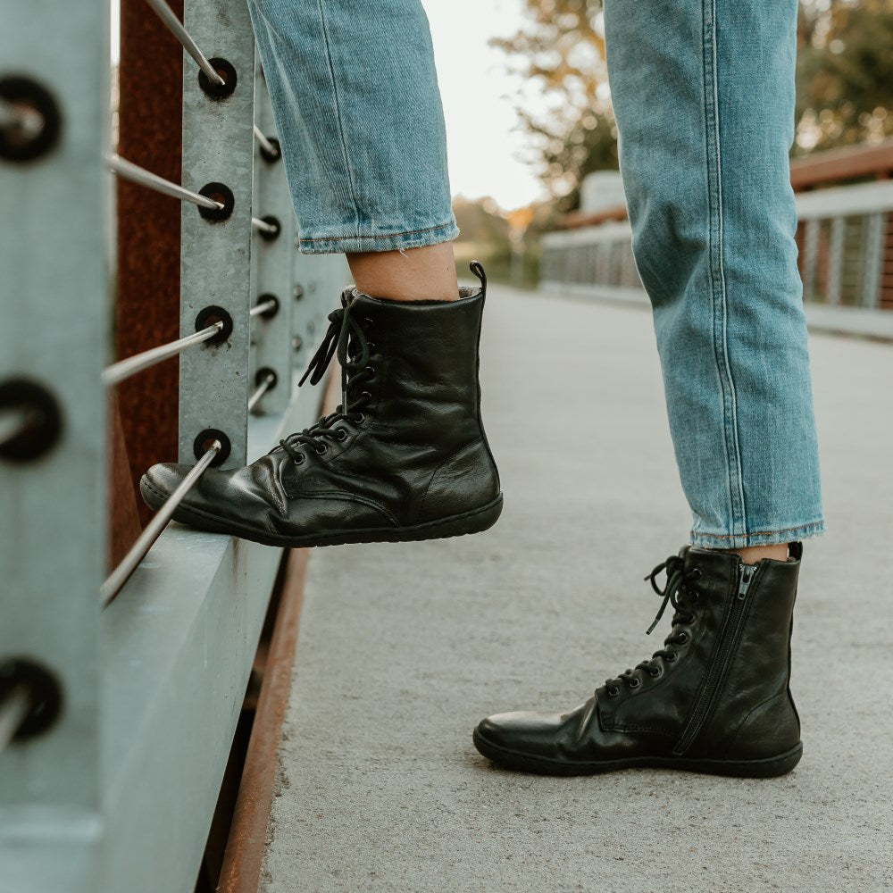 Black, leather, wool-lined Mukishoes Igneous combat-style, zip-up boots. Both boots are shown here facing left on a woman wearing loose, cropped, light-wash jeans, standing on a cement and metal bridge. Left foot is resting on the bottom metal slat of the bridge wall.  #color_black