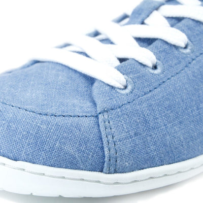 A photo of Mukishoes Indigo fabric sneakers. Shoes are simple in design, have a pull tab on the back, and white lining, laces, and soles. Left shoe is shown here with a close-up of the laces and fabric against a white background. #color_indigo