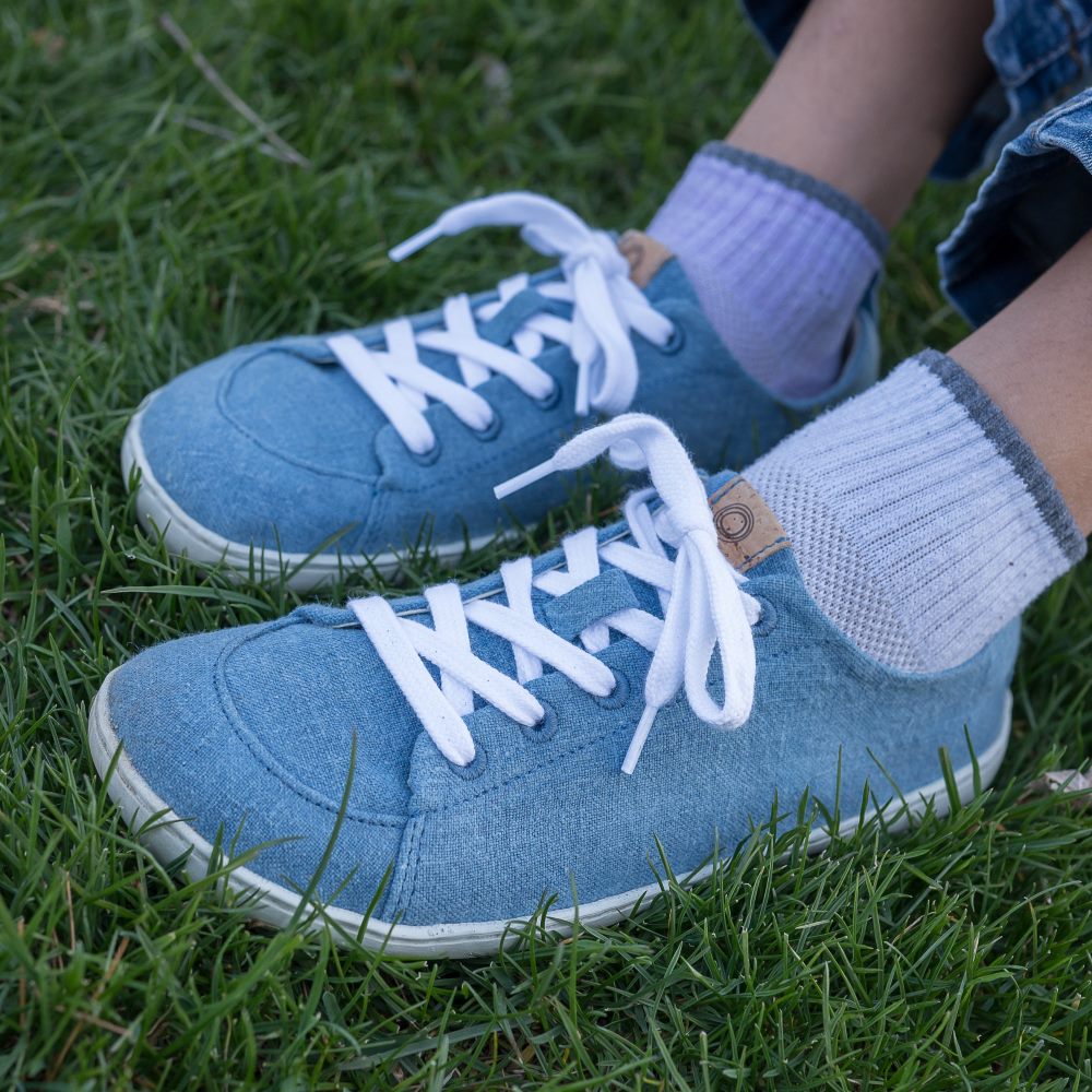 A photo of Mukishoes Indigo fabric sneakers. Shoes are simple in design, have a pull tab on the back, and white lining, laces, and soles. Both shoes are shown here diagonally to the left on a girl sitting in grass. #color_indigo