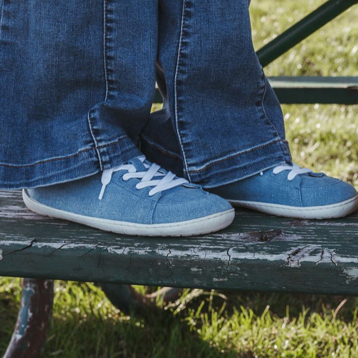A photo of Mukishoes Indigo fabric sneakers. Shoes are simple in design, have a pull tab on the back, and white lining, laces, and soles. Both shoes are shown here facing to the right on a young girl wearing bell bottom jeans standing on a metal and wooden picnic bench. #color_indigo