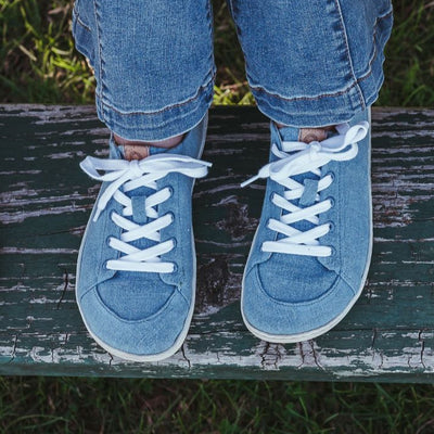 A photo of Mukishoes Indigo fabric sneakers. Shoes are simple in design, have a pull tab on the back, and white lining, laces, and soles. Both shoes are shown here from above on a young girl wearing bell bottom jeans sitting on a metal and wooden picnic bench. #color_indigo