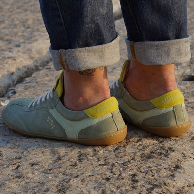 Theia (multiple green colors) Mukishoes Lyra suede leather sneakers with, white laces, and a natural rubber colored sole. Shoes are all sage green colored with a T-shaped detail on the toe box in sage green, a T-shaped seafoam green detail on both sides of the ankle, and neon green color detail on the top of the tongue and heel. Both shoes are shown facing diagonally to the back left on a tan woman wearing rolled, dark jeans standing on pavement. #color_theia