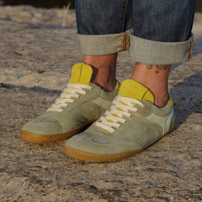 Theia (multiple green colors) Mukishoes Lyra suede leather sneakers with, white laces, and a natural rubber colored sole. Shoes are all sage green colored with a T-shaped detail on the toe box in sage green, a T-shaped seafoam green detail on both sides of the ankle, and neon green color detail on the top of the tongue and heel. Both shoes are shown facing diagonally left on a tan woman wearing rolled, dark jeans standing on pavement. #color_theia