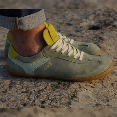 Theia (multiple green colors) Mukishoes Lyra suede leather sneakers with, white laces, and a natural rubber colored sole. Shoes are all sage green colored with a T-shaped detail on the toe box in sage green, a T-shaped seafoam green detail on both sides of the ankle, and neon green color detail on the top of the tongue and heel. Both shoes are shown facing right on a tan woman wearing rolled, dark jeans standing on pavement. #color_theia