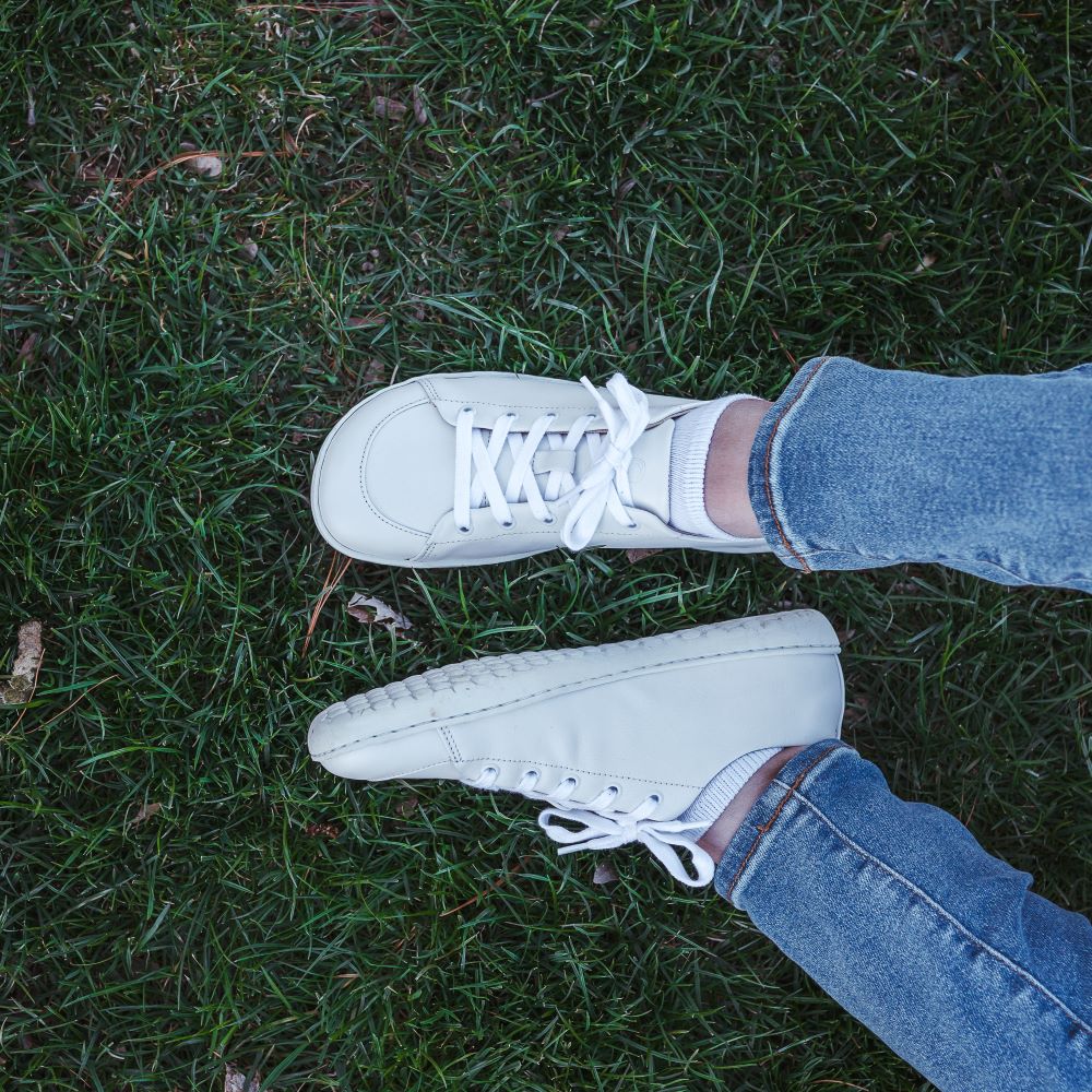 A photo of white Mukishoes Raw Leather Cloud sneakers. Shoes are simple in design, have a pull tab on the back, and tan leather lining. Left shoe is shown here leaning to the left while right shoe is shown from above on a tan woman wearing medium-wash cropped jeans sitting in grass. #color_raw-leather-cloud