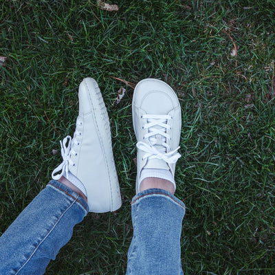 A photo of white Mukishoes Raw Leather Cloud sneakers. Shoes are simple in design, have a pull tab on the back, and tan leather lining. Left shoe is shown here leaning to the left while right shoe is shown from above on a tan woman wearing medium-wash cropped jeans sitting in grass. #color_raw-leather-cloud