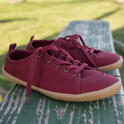 A photo of Mukishoes Roma fabric sneakers. Shoes are a deep wine color, simple in design, have a pull tab on the back, and a deep-wine colored lining, laces, and gum colored soles. Both shoes are shown here diagonally facing right on an old green bench with grass in the distance. #color_roma