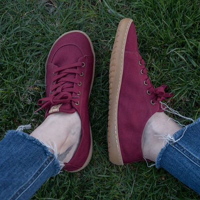 A photo of Mukishoes Roma fabric sneakers. Shoes are a deep wine color, simple in design, have a pull tab on the back, and a deep-wine colored lining, laces, and gum colored soles. Left shoe is shown here leaning to the left while right shoe is shown from above on a tan woman wearing medium-wash cropped jeans sitting in grass. #color_roma