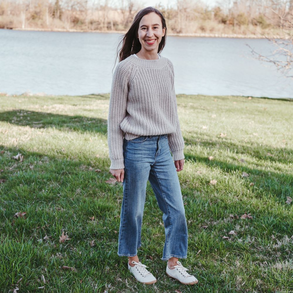 A photo of Mukishoes Sand fabric sneakers. Shoes are simple in design with a subtle herringbone pattern, brass-colored rivets, a pull tab on the back, and sand-colored lining, laces, and gum-colored soles. Both shoes are shown diagonally to the right here on a brown-haired woman wearing a cream, chunky sweater and light-wash wide-legged crop jeans standing on grass with a small lake in the distance. #color_sand