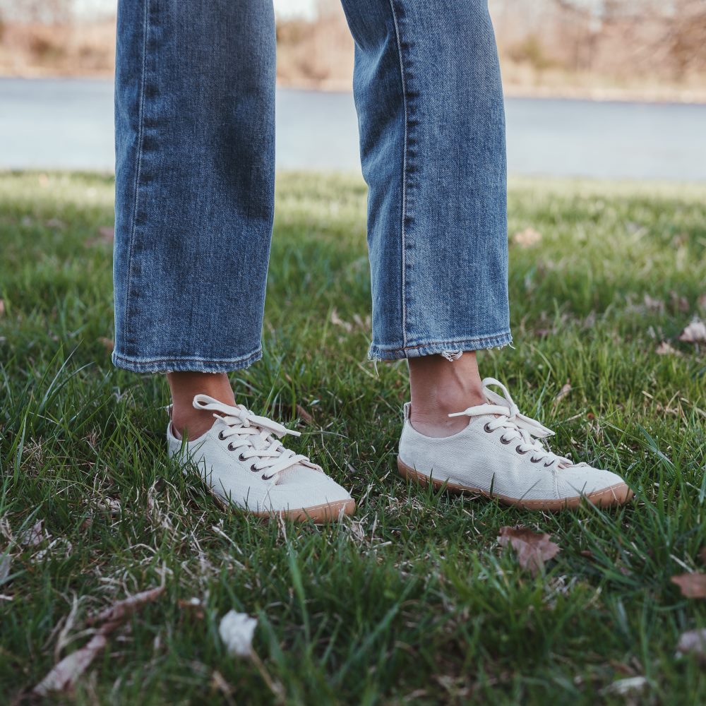 A photo of Mukishoes Sand fabric sneakers. Shoes are simple in design with a subtle herringbone pattern, brass-colored rivets, a pull tab on the back, and sand-colored lining, laces, and gum-colored soles. Both shoes are shown diagonally to the right here on a woman wearing light-wash, wide-legged crop jeans standing on grass with a small lake in the distance. #color_sand