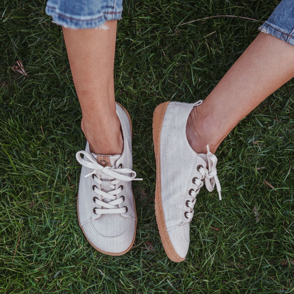 A photo of Mukishoes Sand fabric sneakers. Shoes are simple in design with a subtle herringbone pattern, brass-colored rivets, a pull tab on the back, and sand-colored lining, laces, and gum-colored soles. Left shoe is shown here leaning to the left while right shoe is shown from above on a tan woman wearing medium-wash cropped jeans sitting in grass.. #color_sand