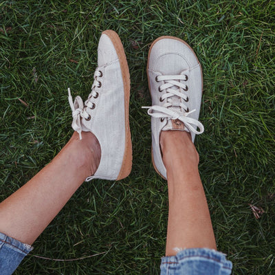 A photo of Mukishoes Sand fabric sneakers. Shoes are simple in design with a subtle herringbone pattern, brass-colored rivets, a pull tab on the back, and sand-colored lining, laces, and gum-colored soles. Left shoe is shown here leaning to the left while right shoe is shown from above on a tan woman wearing medium-wash cropped jeans sitting in grass.. #color_sand