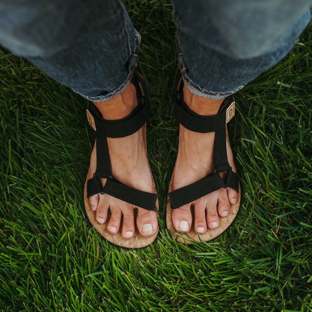 Black Mukishoes Solstice. Sandals have thicker cotton adjustable straps going over the toes and surrounding the ankle with straps connecting the toe and ankle straps. Footbed is quark and sole is a thin black rubber. Both shoes are shown from above on a tan woman wearing rolled jeans standing in grass. #color_black