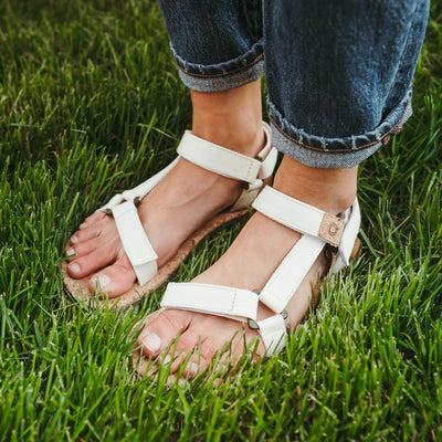 Cream Mukishoes Solstice. Sandals have thicker cotton adjustable straps going over the toes and surrounding the ankle with straps connecting the toe and ankle straps. Footbed is quark and sole is a thin tan rubber. Both shoes are facing diagonally left on a tan woman wearing rolled jeans standing in grass. #color_natural-cream