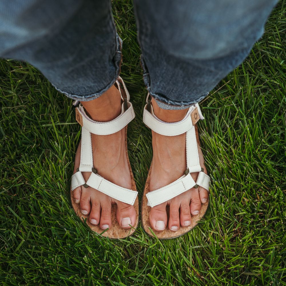 Cream Mukishoes Solstice. Sandals have thicker cotton adjustable straps going over the toes and surrounding the ankle with straps connecting the toe and ankle straps. Footbed is quark and sole is a thin tan rubber. Both shoes are shown from above on a tan woman wearing rolled jeans standing in grass. #color_natural-cream