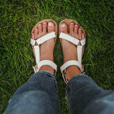 Cream Mukishoes Solstice. Sandals have thicker cotton adjustable straps going over the toes and surrounding the ankle with straps connecting the toe and ankle straps. Footbed is quark and sole is a thin tan rubber. Both shoes are shown from above on a tan woman wearing rolled jeans standing in grass. #color_natural-cream