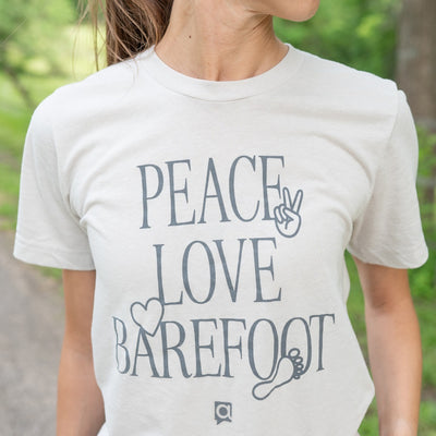 Cool Grey "Peace, Love, Barefoot" T-shirt shown on a tan woman with brown hair with grass and woods in the background.
