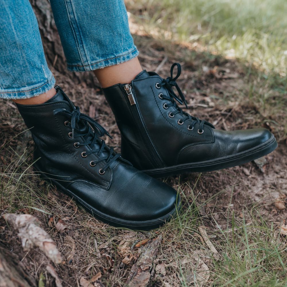 Black Peerko Breeze unlined leather combat boots. Both boots are shown staggered facing right on a person wearing loose-fitting light blue jeans standing in grass close to a tree. #color_black