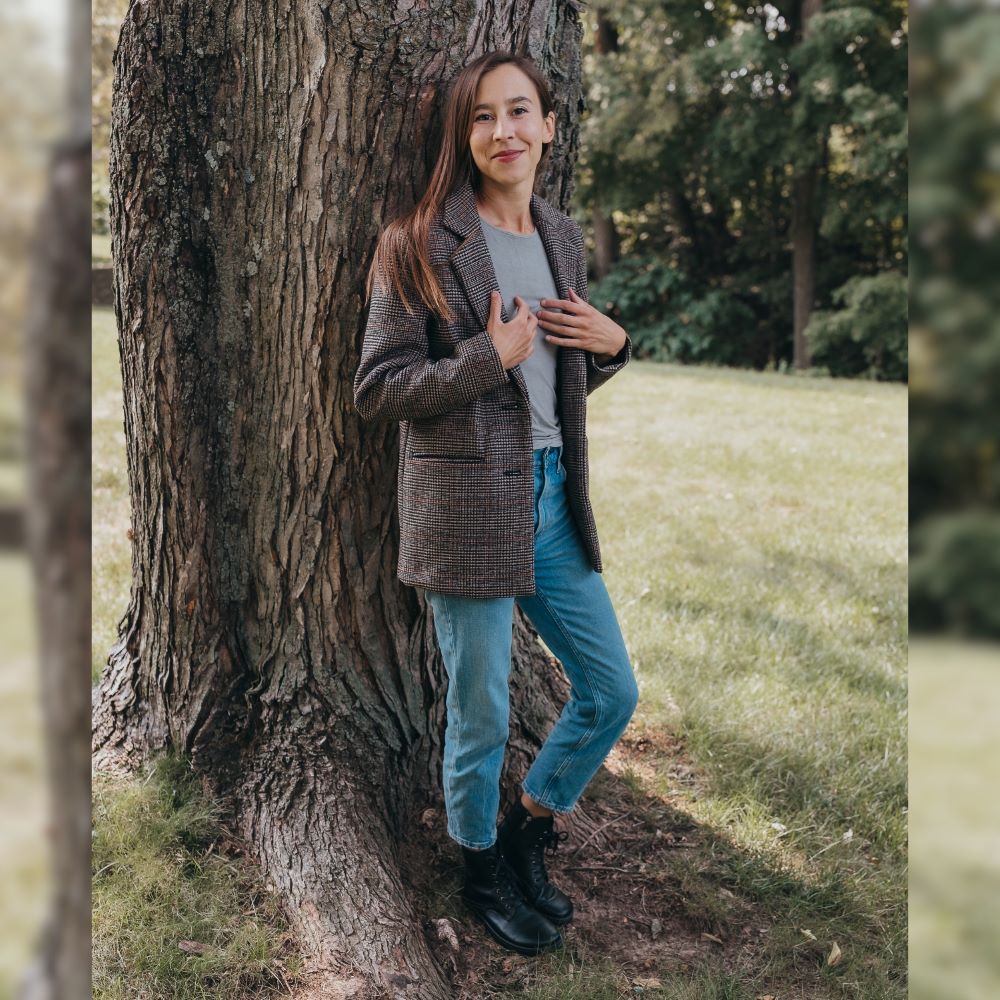 Black Peerko Breeze unlined leather combat boots. Both boots are shown staggered facing right on a woman with dark brown hair wearing loose-fitting light blue jeans, a grey shirt, and a long brown tweed blazer standing in grass close to a tree. #color_black