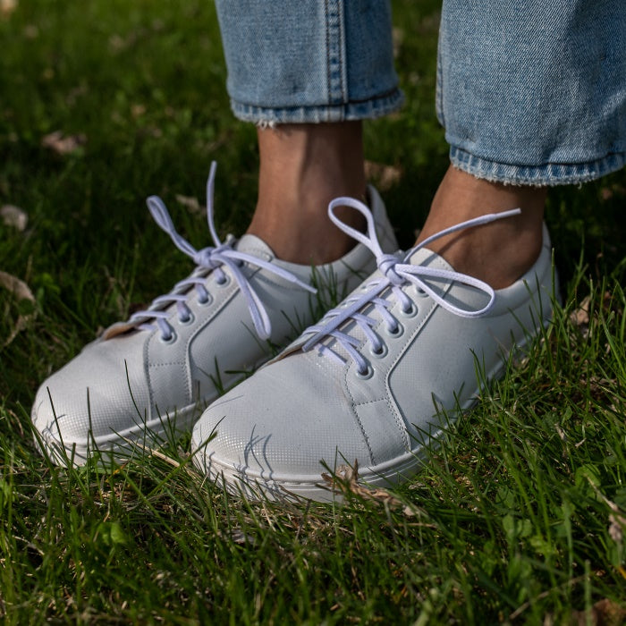 A photo of white Peerko Celebrate sneakers made with leather and recyclable soles. Sneakers have a slight sparkle. Shown diagonally being worn by a woman wearing lightwash ankle height jeans standing in grass. #color_white