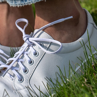 A photo of white Peerko Celebrate sneakers made with leather and recyclable soles. Sneakers have a slight sparkle. Shown as a close up to show details diagonally being worn by a woman wearing lightwash ankle height jeans standing in grass. #color_white