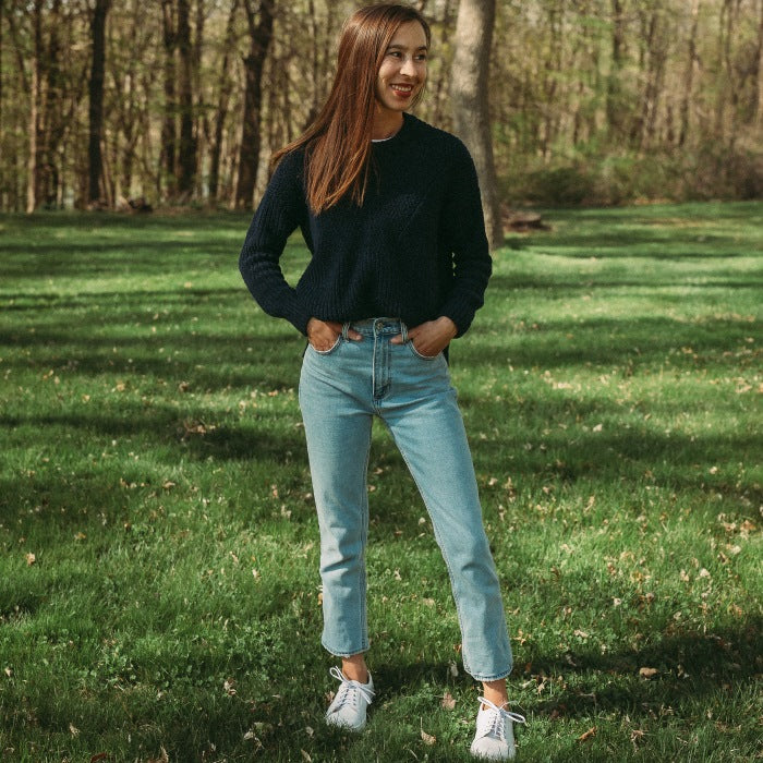 A photo of white Peerko Celebrate sneakers made with leather and recyclable soles. Sneakers have a slight sparkle. Shown from the front being worn by a woman wearing lightwash ankle height jeans and a navy blue sweater standing in grass. #color_white