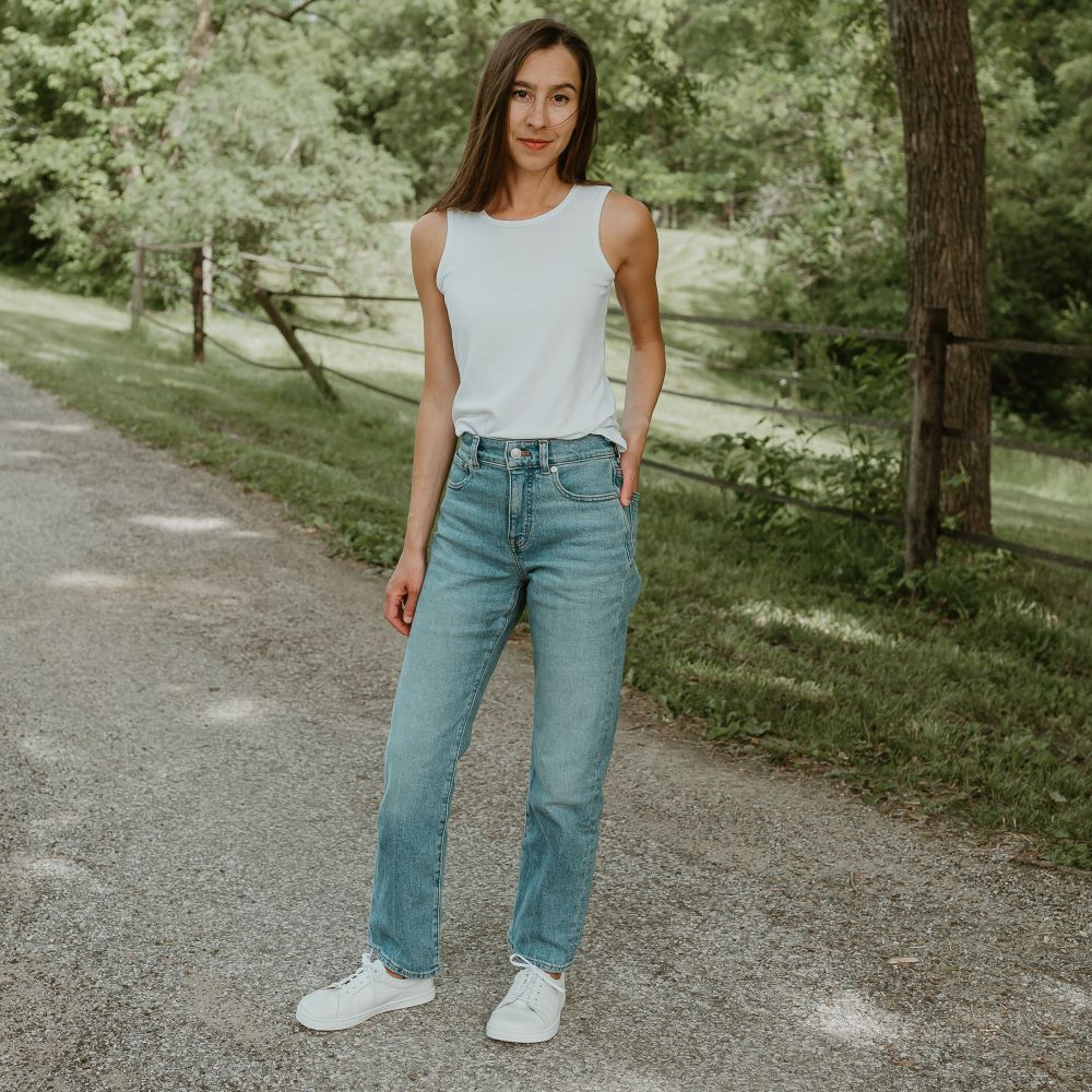 All white Peerko Classic simple leather sneakers. Shoes are shown on a tan woman with long brown hair wearing a white tank top and loose, light-wash blue-jeans. Her right hand is in her back right pocket and her left foot is turned to the side. She is on a cement road with a fance, grass, and woods in the background. #color_white