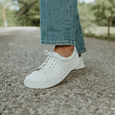 All white Peerko Classic simple leather sneakers. Shoes are shown facing diagonally to the left on a tan woman wearing loose, light-wash blue-jeans standing on a cement road with a fence, grass, and woods in the background. #color_white