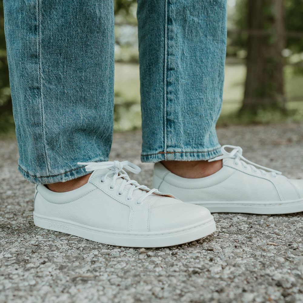 All white Peerko Classic simple leather sneakers. Shoes are facing to the right on a tan woman wearing loose, light-wash blue-jeans standing on a cement road with a fence, grass, and woods in the background. #color_white