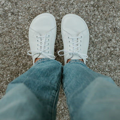 All white Peerko Classic simple leather sneakers. Shoes are shown from above on a tan woman wearing loose, light-wash blue-jeans standing on a cement. #color_white